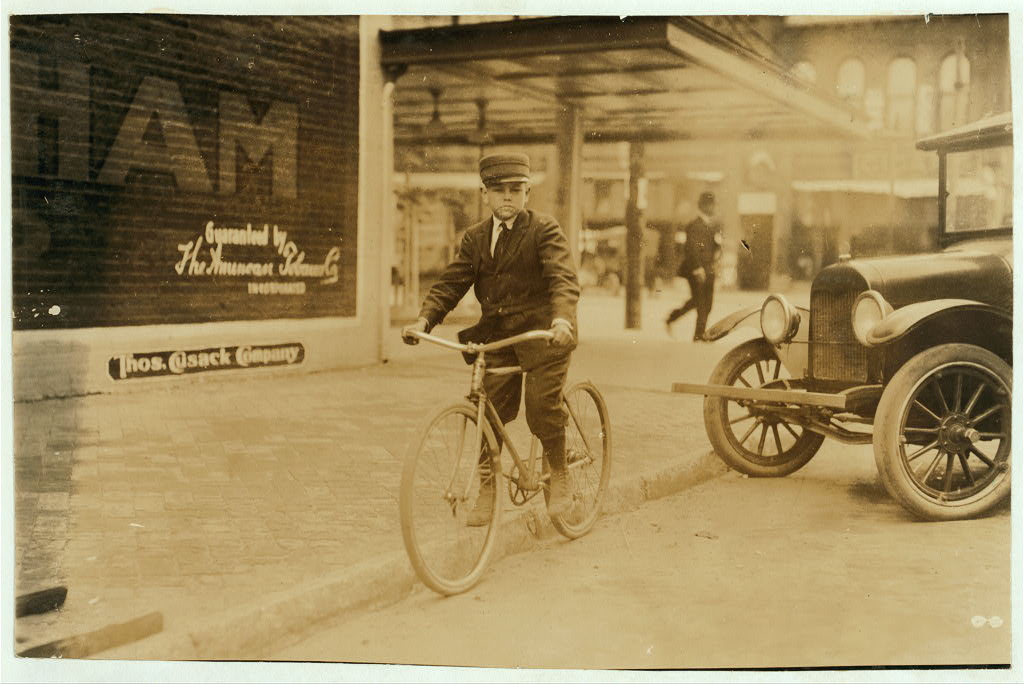 A young messenger boy on a bicycle beside an early automobile on a brick-paved downtown street, Oklahoma City, c. early 1900s